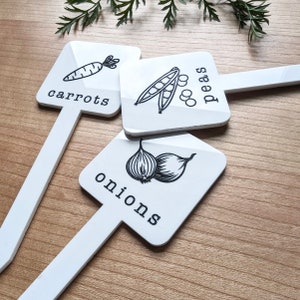 May include: Three white garden markers with black illustrations and text. The markers are shaped like a rectangle with rounded corners. The text on the markers reads "carrots", "peas", and "onions".