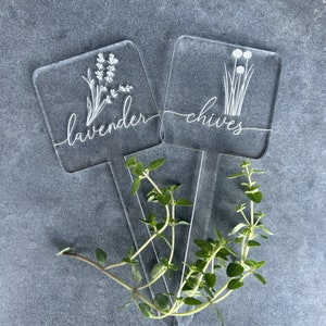 May include: Two clear acrylic plant markers with white lettering and illustrations. The marker on the left says "lavender" and has a drawing of lavender flowers. The marker on the right says "chives" and has a drawing of chives.