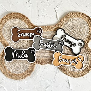 K&ouml;nnte beinhalten: F&uuml;nf h&ouml;lzerne, knochenf&ouml;rmige Hundeanh&auml;nger mit wei&szlig; bemalten Kanten. Die Anh&auml;nger sind mit den Namen "Snoopy", "Buster", "Moose", "Mila" und "Bouswer" personalisiert. Jeder Anh&auml;nger hat ein Pfotenabdruck-Design.