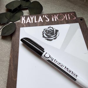 May include: A wooden dry erase board with the text "KAYLA'S NOTES" in rose gold lettering. A black and white rose graphic is on the white surface. A black and white dry erase marker is in the foreground.