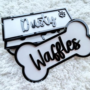 Könnte beinhalten: Zwei weiße Hundename-Schilder mit schwarzen Rändern. Ein Schild ist ein Rechteck mit dem Namen "Dusty" und einem Pfotenabdruck. Das andere Schild ist knochenförmig mit dem Namen "Waffles".