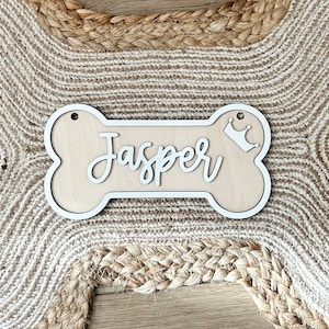 K&ouml;nnte beinhalten: Wei&szlig;er Holz-Hunde-Knochen mit dem Namen "Jasper" in Kursivschrift und einer kleinen Krone auf der rechten Seite.