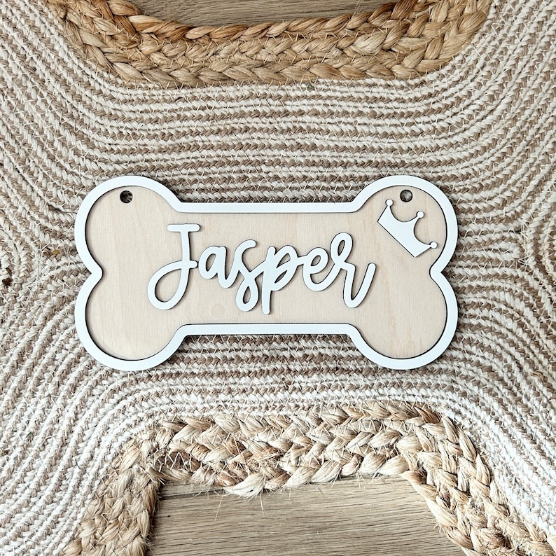 K&ouml;nnte beinhalten: Wei&szlig;er Holz-Hunde-Knochen mit dem Namen "Jasper" in Kursivschrift und einer kleinen Krone auf der rechten Seite.