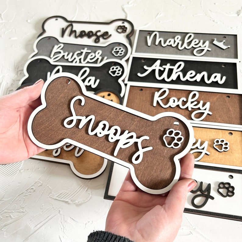 K&ouml;nnte beinhalten: Eine Sammlung von Namensschildern in Form von Hundebeinen aus Holz mit wei&szlig;en Buchstaben. Die Namen auf den Schildern sind "Moose", "Buster", "Lola", "Snoopy", "Marley", "Athena", "Rocky" und "Lucy".