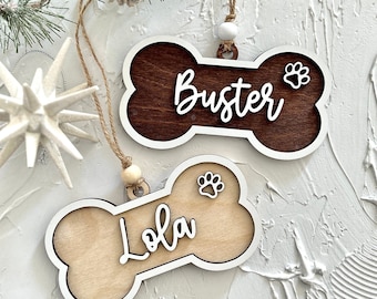 Può includere: Due decorazioni in legno a forma di osso di cane con impronte di zampe bianche e i nomi "Buster" e "Lola" scritti in corsivo bianco. Le decorazioni sono appese a uno spago con perline bianche.