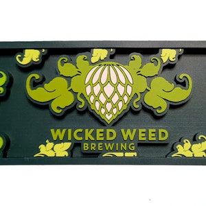 May include: A rectangular dark gray sign featuring the text WICKED WEED BREWING in raised lettering. The design includes a central stylized hop cone graphic in light green and white, surrounded by decorative green leafy scrollwork patterns.