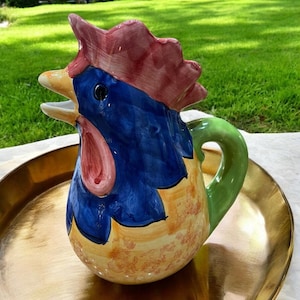 May include: A ceramic rooster pitcher with a green handle. The rooster is painted in shades of blue, yellow, red, and orange.