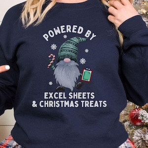 May include: Navy blue sweatshirt with a Christmas gnome graphic. The gnome holds a candy cane and a clipboard, with the text "POWERED BY EXCEL SHEETS & CHRISTMAS TREATS". The sweatshirt is a crew neck style.