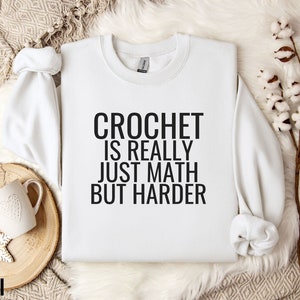 May include: White sweatshirt with black text that reads "Crochet is really just math but harder".