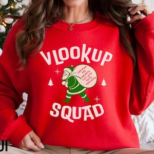 Może przedstawiać: Czerwona bluza z białym napisem "VLOOKUP SQUAD". Zielona grafika Świętego Mikołaja niesie worek z napisem "Checking the Naughty List with Excel Precision". Na bluzie znajdują się również dwa małe białe drzewka bożonarodzeniowe.