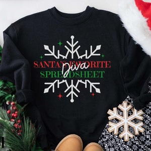 Könnte beinhalten: Schwarzes Sweatshirt mit dem Text "Santa's Favorite Diva Spreadsheet" in Rot und Grün, mit einem weißen Schneeflocken-Design. Das Bild zeigt Weihnachtsdekorationen, darunter einen Weihnachtsmannhut und einen Schneeflocken-Schmuck.