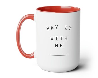 Könnte beinhalten: Weiße Keramiktasse mit rotem Rand und Henkel. Die Tasse hat einen schwarzen Text, der "Say it with me" mit einer Linie darunter lautet.