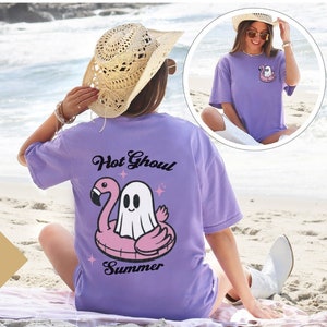 May include: A violet t-shirt featuring a ghost on a pink flamingo floatie, with the words "Hot Ghoul Summer". The shirt is worn on a beach with the sea in the background. A smaller image shows the front of the shirt.