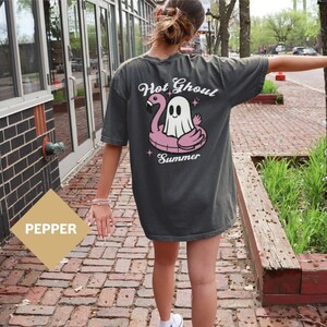May include: A dark gray t-shirt with a graphic of a ghost riding a pink flamingo float. The text "Hot Ghoul Summer" is printed on the shirt.