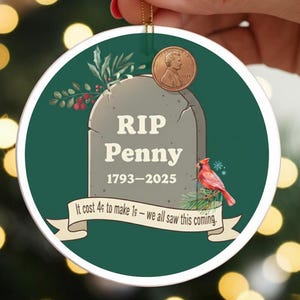 May include: A round Christmas ornament with a green background and a tombstone design that reads "RIP Penny 1793-2025." A penny is affixed to the top. The ornament also features a red bird and the text "It cost 4¢ to make 1¢ - we all saw this coming."