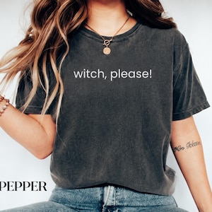 May include: A charcoal grey short-sleeve t-shirt with a crew neckline featuring the white text "witch, please!" printed across the chest. The shirt has a washed-out, vintage-inspired appearance and is paired with blue denim jeans.