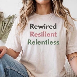 May include: A beige t-shirt featuring the words "Rewired, Resilient, Relentless" in black, pink, and green. The shirt is paired with blue jeans. The design conveys a message of resilience and determination.