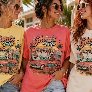 May include: Three women wearing oversized t-shirts with a "Ghouls Trip" graphic. The design features ghosts, a skeleton, a vintage car, and luggage. The shirts are in yellow, coral, and cream colors, paired with denim shorts.
