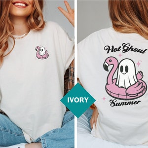 May include: Ivory t-shirt with a cartoon ghost on a pink flamingo inflatable. The back of the shirt has the words "Hot Ghoul Summer" above the flamingo and ghost design. The t-shirt has a loose fit.