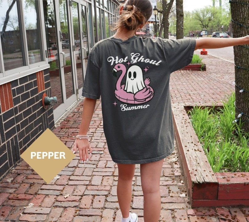 May include: A dark gray t-shirt with a graphic of a ghost riding a pink flamingo float. The text "Hot Ghoul Summer" is printed on the shirt.