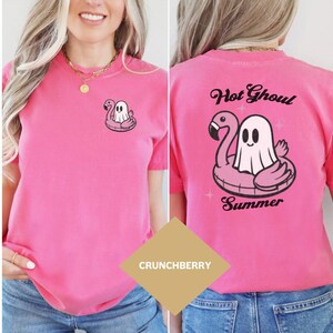 May include: A vibrant pink t-shirt with a cartoon ghost in a flamingo floatie design. The back of the shirt displays the text "Hot Ghoul Summer" above the same graphic. The tee is a solid colour and has short sleeves.