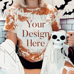 May include: A woman wearing a bleached orange and white tie-dye t-shirt with the text "Your Design Here" on the front. She is holding a white skull mug and wearing a white knitted blanket.