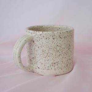 May include: A speckled ceramic mug with a rounded handle. The mug is off-white with small, dark brown speckles. The handle has a ridged design. The mug is set against a soft pink background.