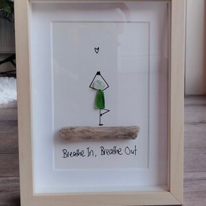 May include: Framed artwork depicting a yoga pose crafted from sea glass and driftwood. The figure is in a tree pose, with a heart above. The text "Breathe In, Breathe Out" is below. The frame is made of light-coloured wood.