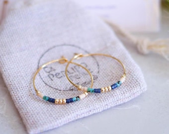 Può includere: Un paio di orecchini a cerchio dorati con piccole perline colorate nei toni del blu, bianco e oro. Gli orecchini poggiano su un piccolo sacchetto di lino beige con chiusura a coulisse. Sullo sfondo una carta con testo dorato.