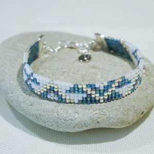 May include: A beaded bracelet with a silver clasp, resting on a grey stone. The bracelet features a pattern of white, blue, and iridescent beads. The bracelet is approximately 2 cm wide.