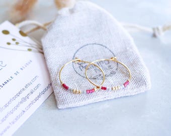 Può includere: Un paio di orecchini a cerchio dorati con piccole perline rosse, bianche e dorate. Gli orecchini sono esposti su un piccolo sacchetto con coulisse bianco sporco. Sullo sfondo è visibile un biglietto da visita con testo.