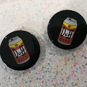 May include: Two black round buttons with a white background and a yellow and orange can of Duff Beer with the text "Duff Beer" on it.