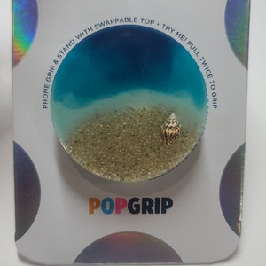 May include: A blue and white phone grip with a seashell design. The grip has a clear, resin-like top with a blue ocean and white sand design. The text "POPGRIP" is printed on the bottom of the grip.