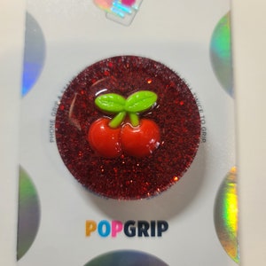 May include: A red glitter PopGrip with a cherry design. The cherry is red with green leaves. The PopGrip has the words "POPGRIP" in colorful letters at the bottom.