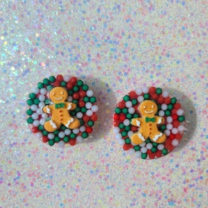 May include: Two round Christmas-themed ornaments. Each ornament features a gingerbread man with a green bow tie, surrounded by red, green, and white beads. The gingerbread men are orange with white accents.