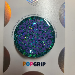 May include: A teal blue PopGrip phone grip with a purple leopard print and glitter. The text "PHONE GRIP & STAND WITH SWAPPABLE TOP" and "TWICE TO GRIP" are printed on the white background.