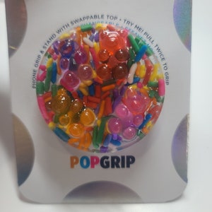 May include: A colorful PopGrip phone accessory with a clear, textured top filled with sprinkles and gel-like shapes in various colors. The packaging reads "PHONE GRIP & STAND WITH SWAPPABLE TOP. TRY ME! PULL TWICE TO GRIP" and "POPGRIP".