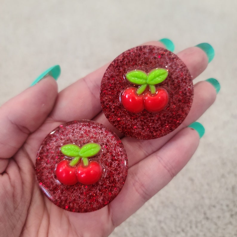 May include: Two red glitter resin cabochons with a green cherry design. Each cabochon features two red cherries with a green stem and leaves.