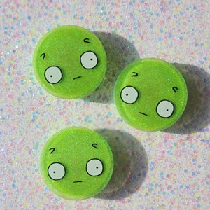 May include: Three green glitter resin buttons with cartoon faces. Each button has two large eyes, a small mouth, and a single eyebrow.