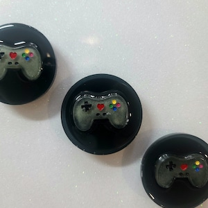 May include: Three black and white round phone grips with a gray video game controller design. Each controller has a red heart, green, yellow, and blue buttons.