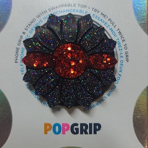 May include: A black and red PopGrip phone accessory in a sunflower design. The center and petals are covered in glitter. The text "POPGRIP" is at the bottom. The product has the text "PHONE GRIP & STAND WITH SWAPPABLE TOP" around the edge.