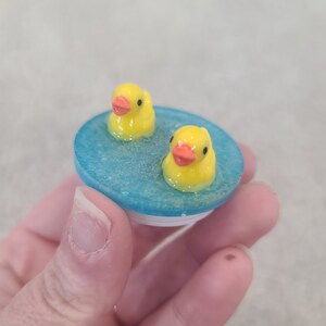 May include: A blue and white phone grip featuring two yellow rubber duckies floating in a blue pool. The ducks have orange beaks and are surrounded by a glittery, textured surface.