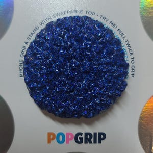 May include: A round, dark blue PopGrip phone accessory with a textured, glittery surface. The accessory is on a white background with the PopGrip logo and text that reads "Phone Grip & Stand with Swappable Top. Try Me! Pull Twice to Grip."