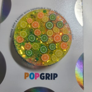May include: A round, clear phone grip with a lime and orange design. The grip has a glittery background and is decorated with lime and orange slices. The text "POPGRIP" is printed on the white background.