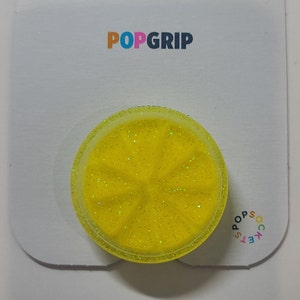 May include: A yellow glitter phone grip shaped like a lemon slice. The grip has a clear outer edge and is designed to be attached to the back of a phone.