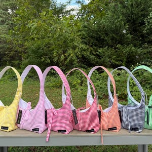 May include: A collection of seven gingham-patterned shoulder bags in various colors: yellow, pink, red, orange, navy, and green. Each bag has a matching handle and a white interior, displayed on a gray table outdoors.