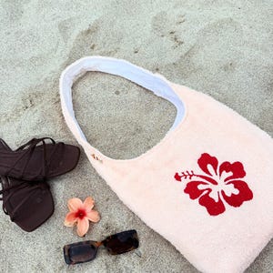 May include: A light pink beach bag with a red hibiscus flower design, brown sandals, and tortoiseshell sunglasses on sand. An orange flower is also present, suggesting a beach or holiday theme.