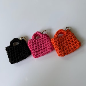 May include: Three small, crocheted bag charms in black, hot pink, and orange. Each bag has a small handle and a silver-colored ring for attachment. The bags are square-shaped and appear to be handmade.