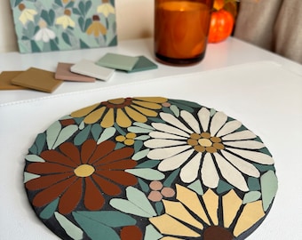 May include: A round mosaic coaster with a floral design. The coaster features a variety of flowers in shades of yellow, white, brown, and green. The flowers are arranged in a circular pattern on a black background.