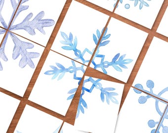May include: Watercolor snowflake designs in shades of blue and light blue are featured on white cards. The cards are arranged on a wooden surface, creating a wintery or holiday theme. The snowflakes have intricate details.